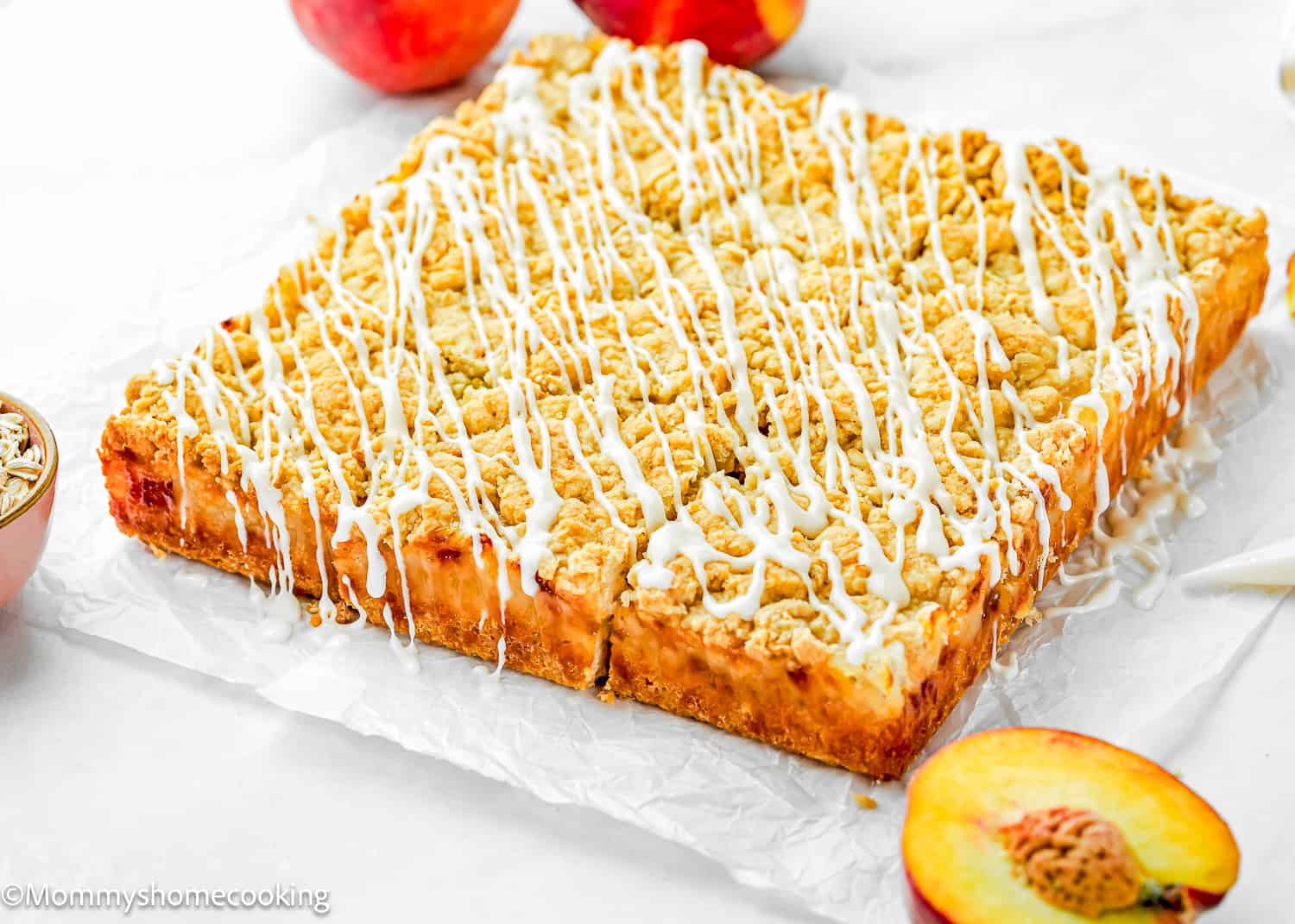 An Eggless Peach Crumb Bar with a crumbly topping and white icing drizzle sits on parchment paper, surrounded by fresh peaches.
