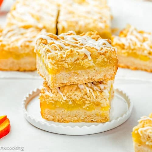 Two Eggless Peach Crumb Bars are stacked on a white plate, topped with a white icing drizzle, with more bars and peach slices in the background.