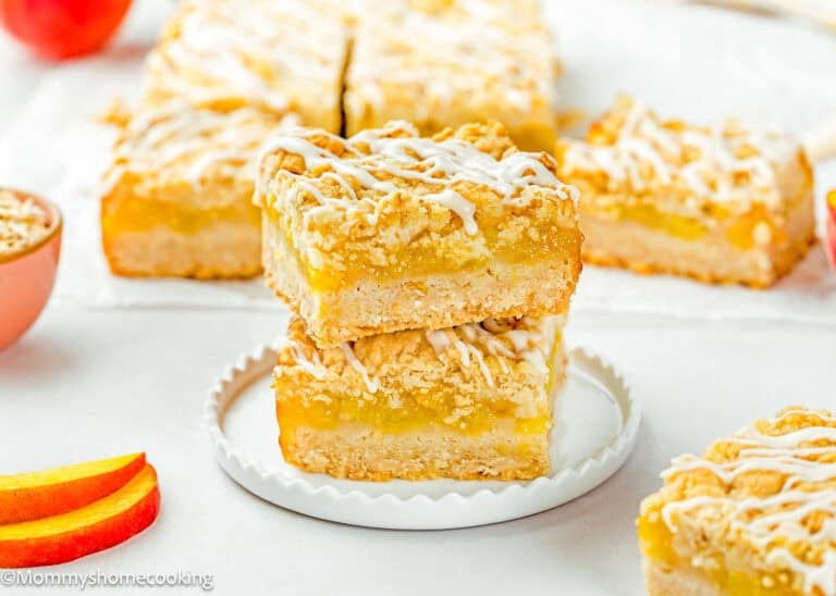 Two Eggless Peach Crumb Bars are stacked on a white plate, topped with a white icing drizzle, with more bars and peach slices in the background.