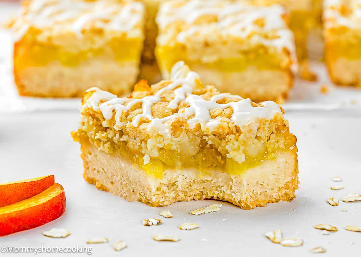 A close-up of an Eggless Peach Crumb Bar with an oat crust, peach filling, crumb topping, and white icing drizzle. Several bars and peach slices are visible in the background.