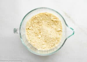 A glass mixing bowl filled with yellow, crumbly flour mixture for Eggless Peach Crumb Bars sits on a white surface.