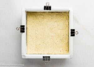 Square baking pan lined with parchment paper and binder clips, filled with an even layer of unbaked dough for Eggless Peach Crumb Bars, on a white surface.