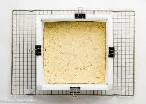 Square baking pan lined with parchment paper and filled with unbaked Eggless Peach Crumb Bars dough, secured with binder clips, sits on a cooling rack.