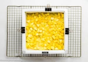 A square baking pan lined with parchment paper, filled with a mixture of diced yellow fruit for Eggless Peach Crumb Bars, sits on a cooling rack.