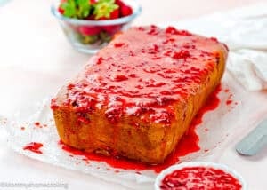 An Eggless Strawberry Pound Cake with Fresh Strawberries sits on parchment paper, covered in a glossy strawberry glaze that drips down the sides. A bowl of strawberries is in the background.