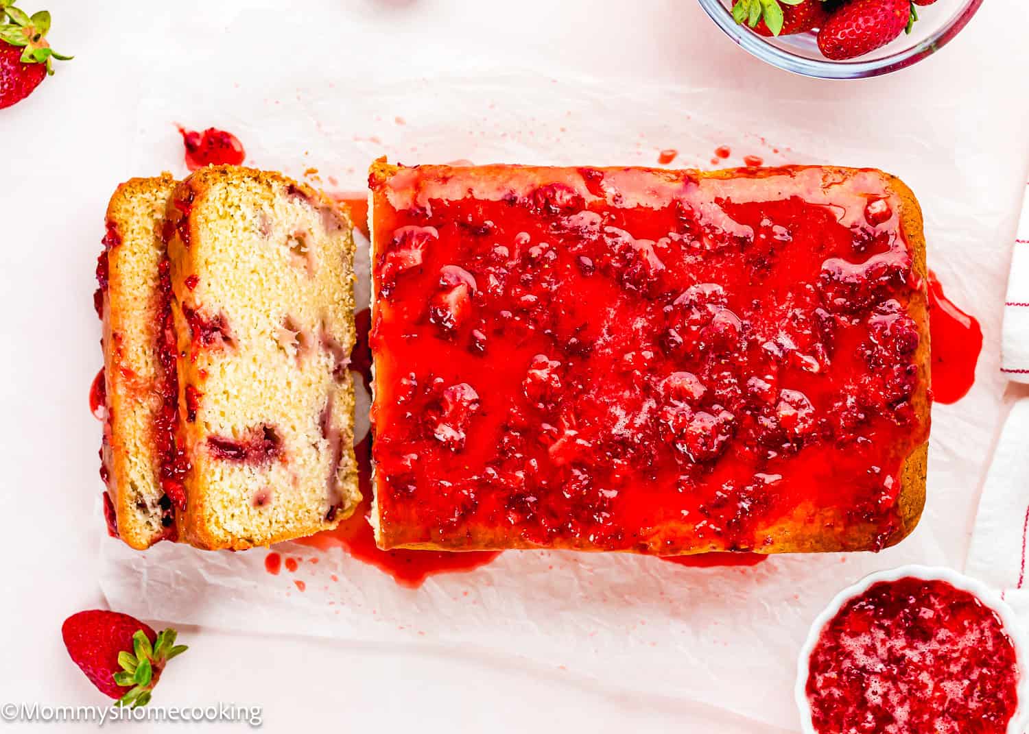 An Eggless Strawberry Pound Cake with Fresh Strawberries is topped with strawberry glaze, partially sliced on parchment paper, surrounded by fresh strawberries and a bowl of glaze.