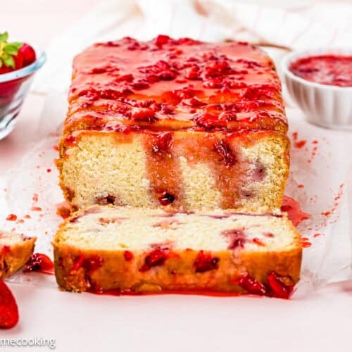 A loaf of Eggless Strawberry Pound Cake with Fresh Strawberries is topped with strawberry glaze and sliced to reveal a moist interior. Fresh strawberries and a bowl of strawberry sauce are placed nearby.