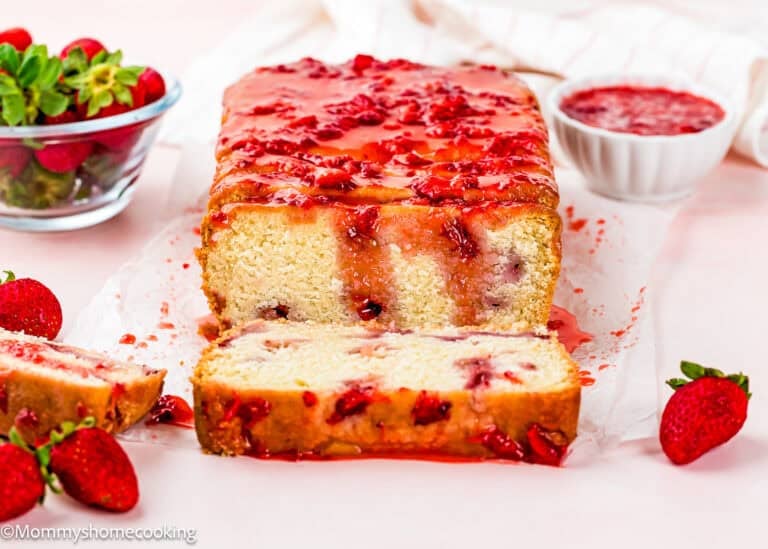 A loaf of Eggless Strawberry Pound Cake with Fresh Strawberries is topped with strawberry glaze and sliced to reveal a moist interior. Fresh strawberries and a bowl of strawberry sauce are placed nearby.