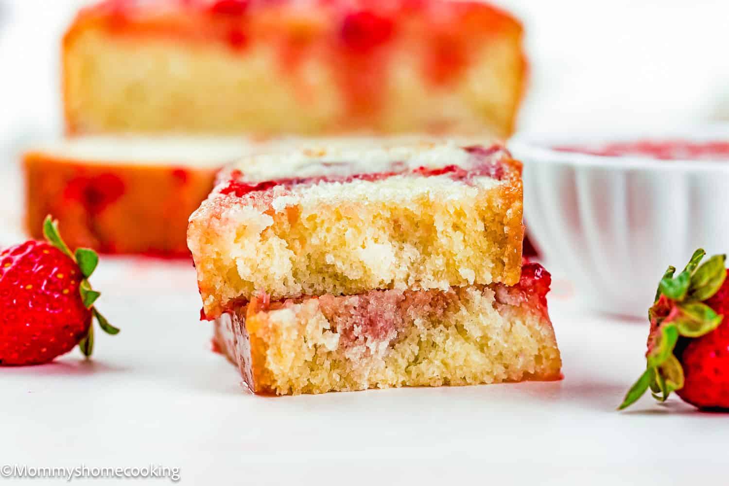 Two slices of Eggless Strawberry Pound Cake with Fresh Strawberries are stacked on a white surface, with fresh strawberries and a bowl in the background.
