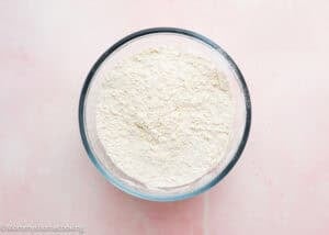 A glass bowl filled with flour sits on a light pink surface, ready for making an Eggless Strawberry Pound Cake with Fresh Strawberries.