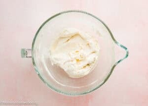 A glass mixing bowl filled with smooth, whipped cream cheese mixture sits on a light pink surface, perfect for making Eggless Strawberry Pound Cake with Fresh Strawberries.