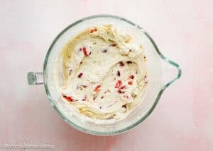 A glass mixing bowl filled with dough for an eggless strawberry pound cake with fresh strawberries, featuring visible pieces of chopped strawberries, placed on a light pink surface.