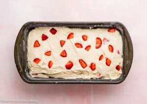 Rectangular loaf pan filled with unbaked batter for Eggless Strawberry Pound Cake with Fresh Strawberries, topped with strawberry pieces on a light pink surface.