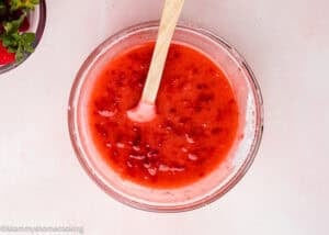 A glass bowl filled with strawberry sauce for Eggless Strawberry Pound Cake with Fresh Strawberries, being mixed with a wooden spoon on a light pink surface.