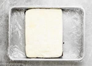 A rectangular block of ice ceram sits on top of Eggless Brownie Ice Cream Sandwiches in a plastic wrap-lined metal baking pan.