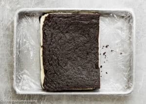 An Eggless Brownie Ice Cream Sandwich rests in a plastic wrap-lined metal baking pan on a gray surface, featuring a rectangular chocolate cake layer with cream filling.
