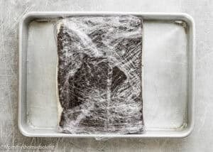A rectangular slab of dough for Eggless Brownie Ice Cream Sandwiches sits on a metal baking tray, wrapped in plastic wrap.