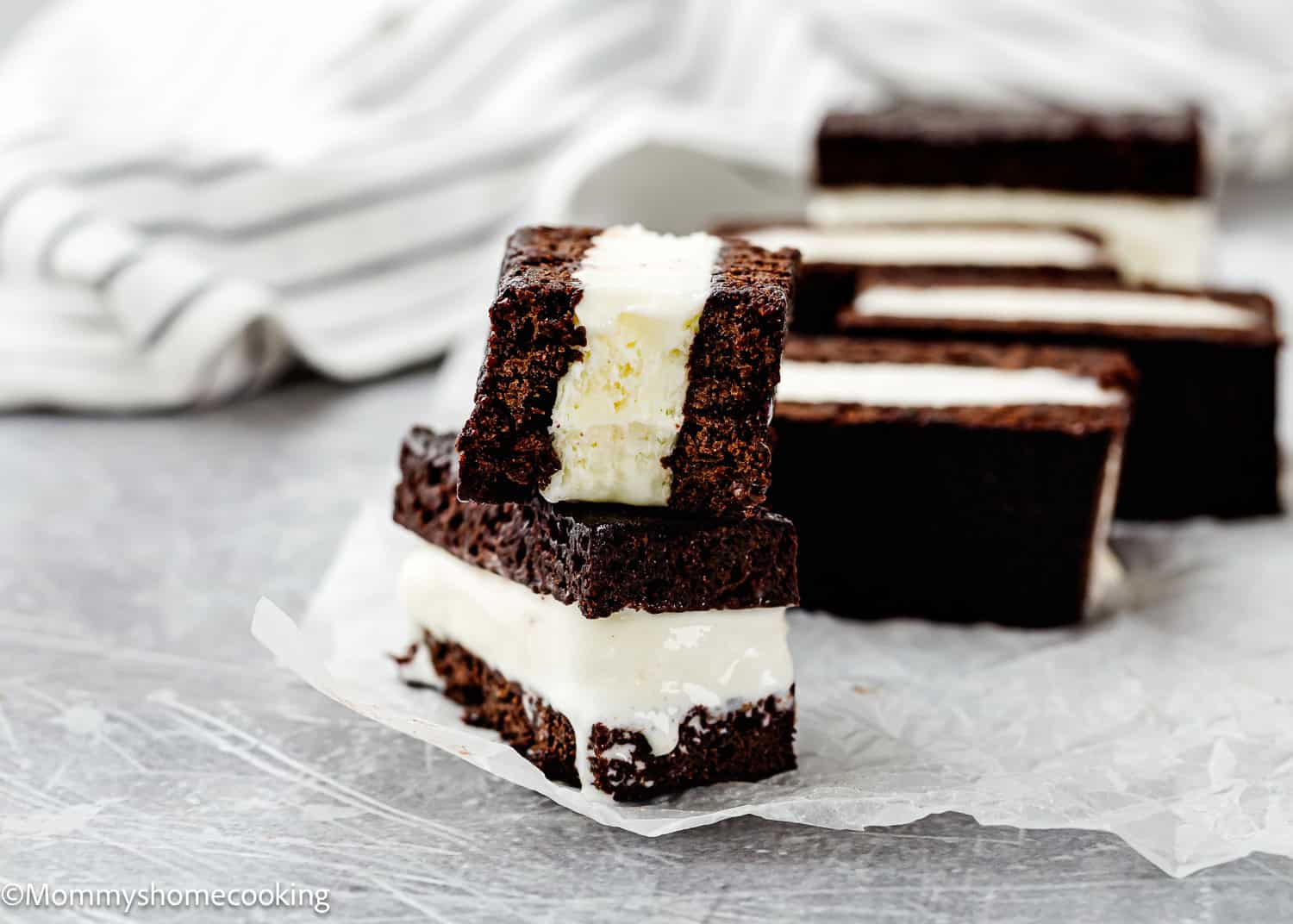 Three Eggless Brownie Ice Cream Sandwiches are stacked on parchment paper, with one sandwich leaning against the others. A striped cloth is visible in the background.