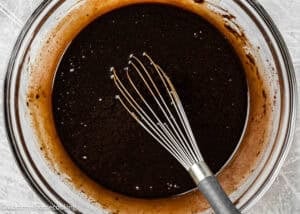 A metal whisk rests in a glass bowl filled with dark chocolate batter for eggless brownie ice cream sandwiches on a light-colored surface.