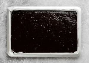 A rectangular baking pan lined with parchment paper filled with unbaked chocolate cake batter for Eggless Brownie Ice Cream Sandwiches on a gray surface.