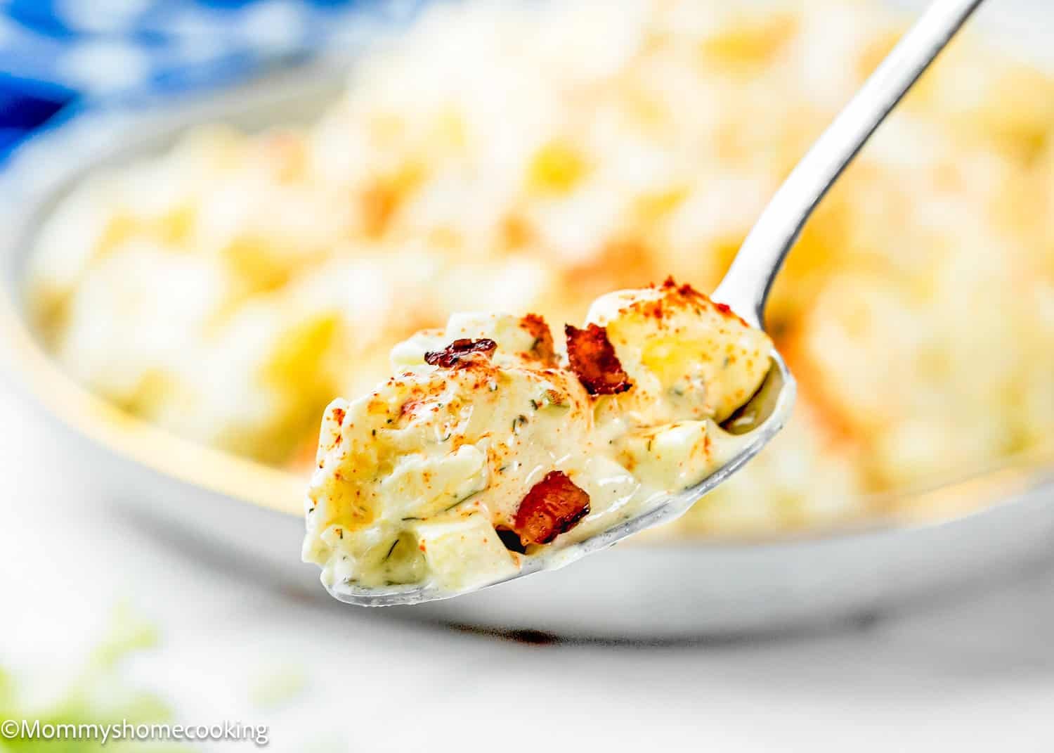 A close-up of a spoonful of Egg-Free Potato Salad (Creamy Classic Recipe) topped with bacon bits, held above a bowl filled with more creamy potato salad.