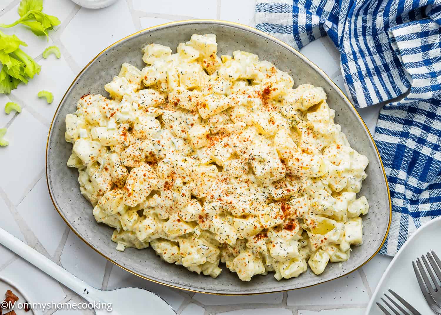 Oval platter of creamy Egg-Free Potato Salad (Creamy Classic Recipe) garnished with paprika and herbs, placed on a white tiled surface with a blue-striped napkin and utensils nearby.