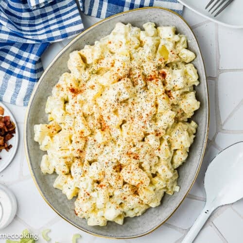 A plate of Egg-Free Potato Salad (Creamy Classic Recipe) topped with paprika sits next to a bowl of bacon pieces, a salt dish, and a large serving spoon on a white tiled surface.