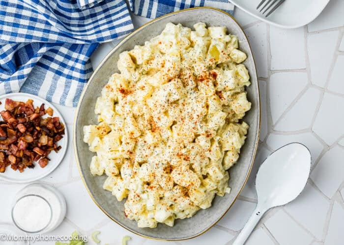 A plate of Egg-Free Potato Salad (Creamy Classic Recipe) topped with paprika sits next to a bowl of bacon pieces, a salt dish, and a large serving spoon on a white tiled surface.