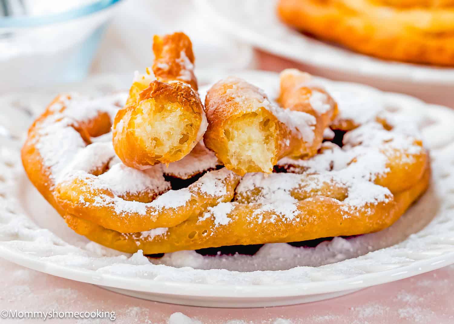 An eggless funnel cake dusted with powdered sugar, topped with pieces of fried dough, served on a white plate.