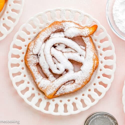 An Eggless Funnel Cake dusted with powdered sugar on a decorative white plate, with a bowl of powdered sugar and a sifter nearby.