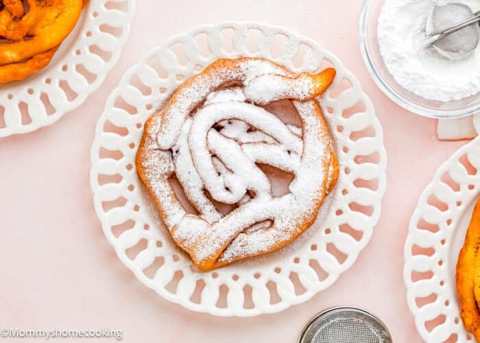 An Eggless Funnel Cake dusted with powdered sugar on a decorative white plate, with a bowl of powdered sugar and a sifter nearby.