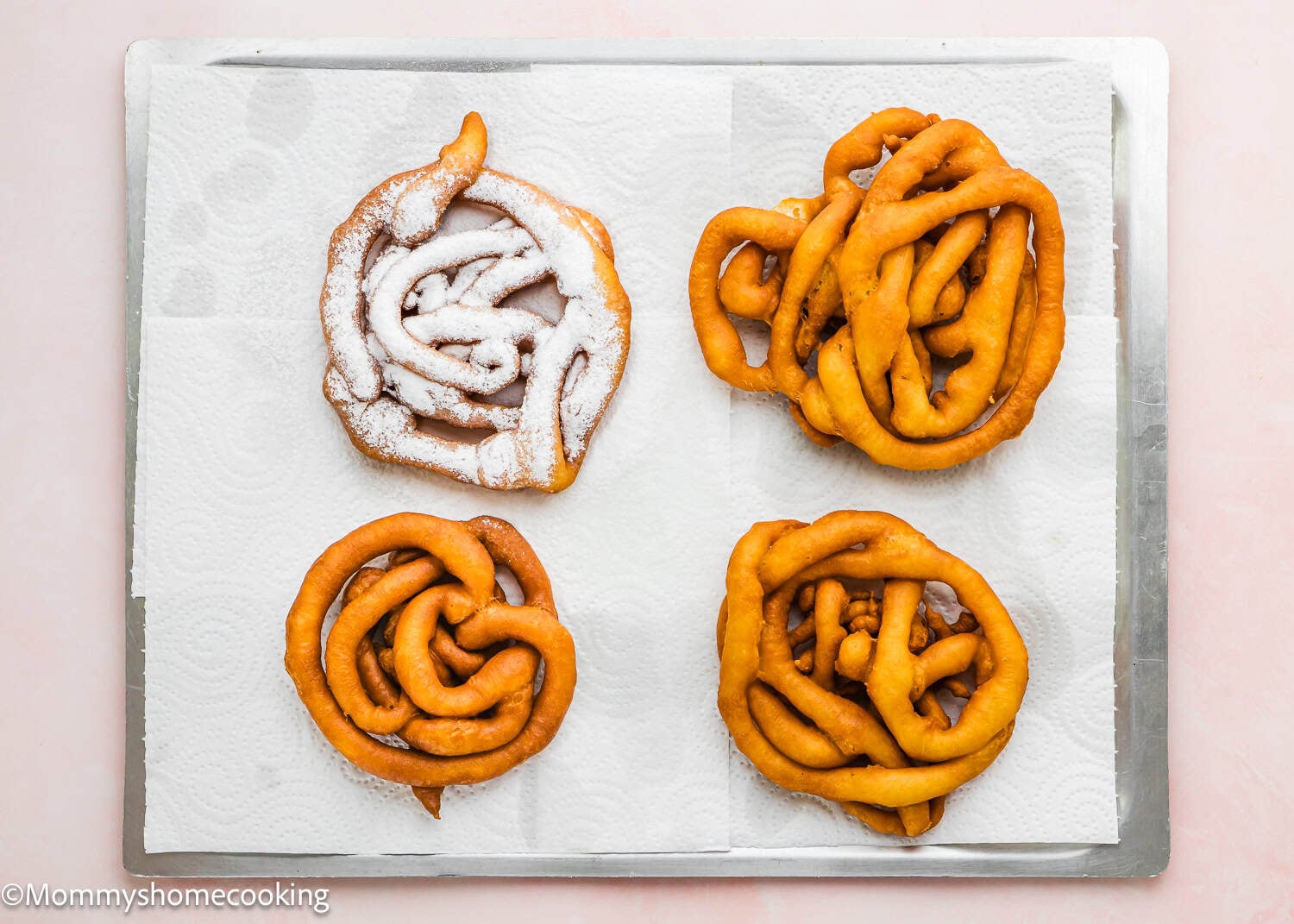 Four pieces of fried funnel cake from an eggless funnel cake recipe rest on paper towels atop a metal baking tray.