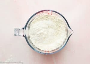 A glass measuring cup filled with flour, ideal for preparing an Eggless Funnel Cake Recipe, sits on a light pink surface.