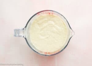 A glass measuring cup filled with thick, creamy batter for an eggless funnel cake recipe sits on a light pink surface.