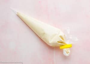 A clear piping bag filled with light-colored cream or frosting, sealed with a yellow clip, sits on a light pink surface—perfect for decorating your Eggless Funnel Cake Recipe.