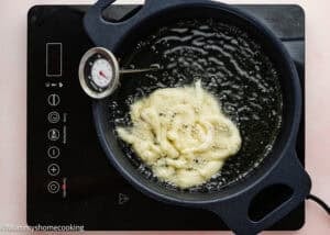 A frying pan with oil and dough being fried for an Eggless Funnel Cake Recipe, a thermometer measures the temperature, all on an electric stovetop.