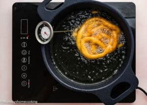 An Eggless Funnel Cake Recipe is being deep-fried in oil in a pan on an electric stovetop, with a thermometer measuring the oil temperature.