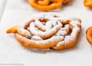A piece of funnel cake from an Eggless Funnel Cake Recipe is dusted with powdered sugar and sits on a white paper towel.