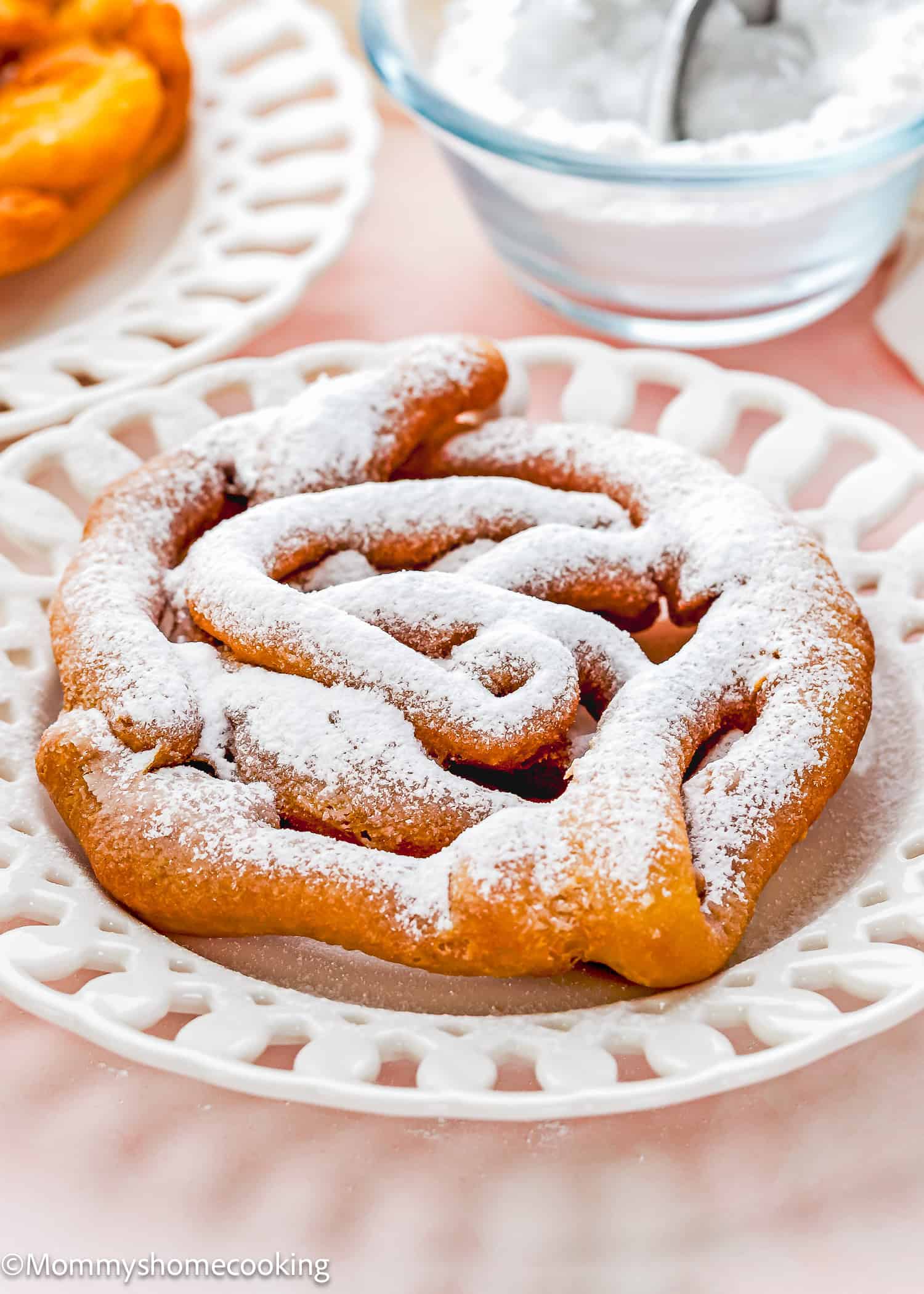 An eggless funnel cake dusted with powdered sugar sits on a white decorative plate, with a bowl of powdered sugar in the background.