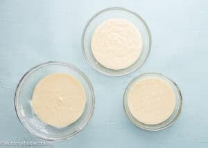 Three clear glass bowls filled with creamy, pale yellow batter are arranged on a light blue surface, perfect for preparing an Eggless Neapolitan Loaf Cake.