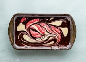 A metal pan filled with chocolate batter swirled with red and white mixtures for an Eggless Neapolitan Loaf Cake, ready for baking, placed on a light blue surface.