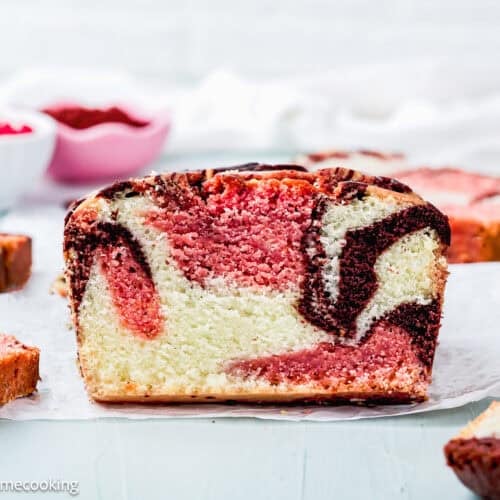 A close-up of a sliced Eggless Neapolitan Loaf Cake with chocolate and strawberry swirls, displayed on parchment paper with more slices in the background.