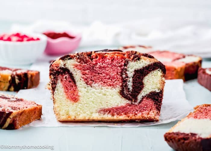 A close-up of a sliced Eggless Neapolitan Loaf Cake with chocolate and strawberry swirls, displayed on parchment paper with more slices in the background.