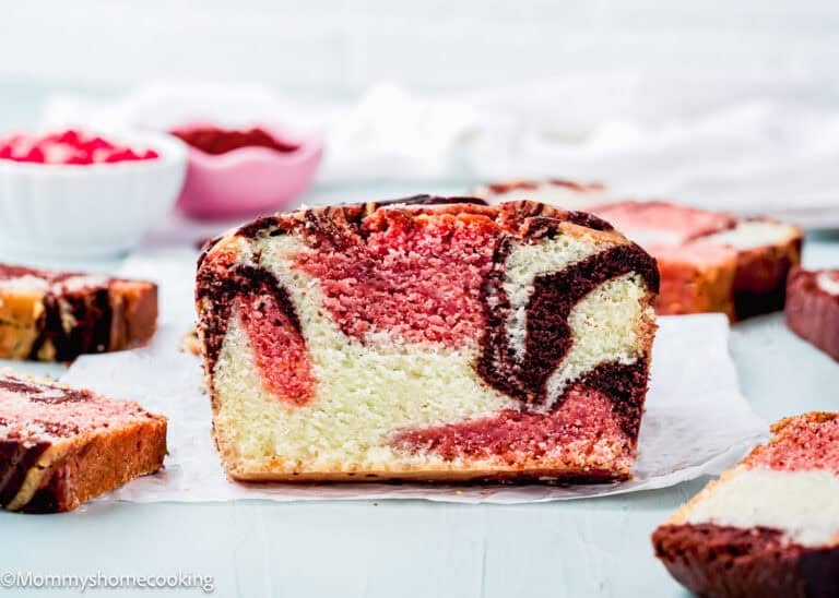 A close-up of a sliced Eggless Neapolitan Loaf Cake with chocolate and strawberry swirls, displayed on parchment paper with more slices in the background.