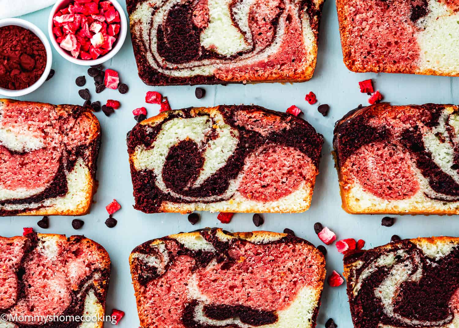Slices of marbled chocolate and strawberry pound cake are arranged on a white surface with chocolate chips and dried strawberry pieces scattered around, showcasing an Eggless Neapolitan Loaf Cake.