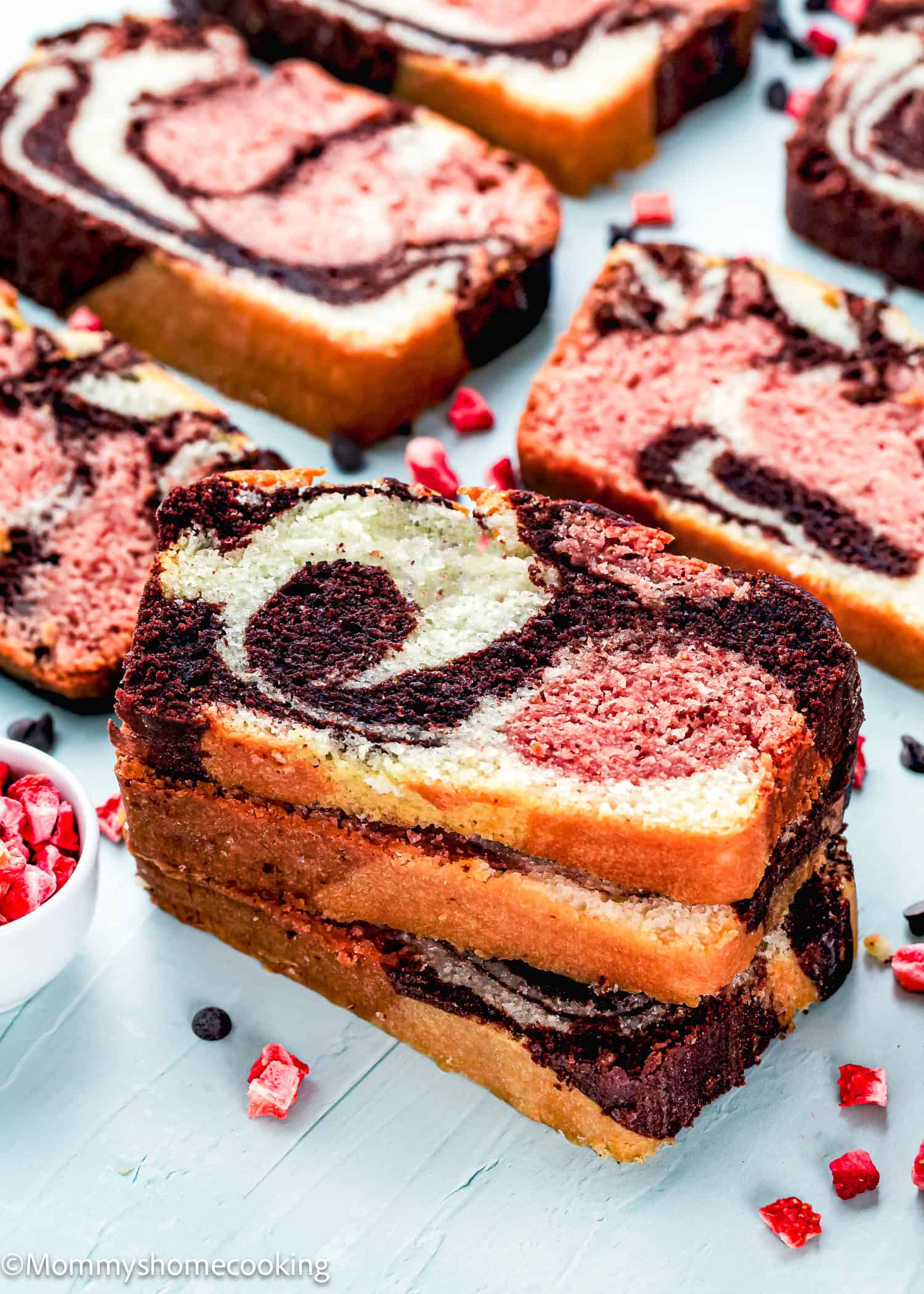 Slices of Eggless Neapolitan Loaf Cake with chocolate and vanilla swirls are stacked and arranged on a white surface, surrounded by small pieces of dried fruit.