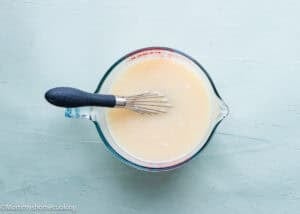 A glass measuring cup filled with pale liquid batter for Eggless Neapolitan Loaf Cake sits on a light blue surface, with a metal whisk resting inside.