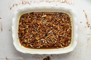 A white baking dish filled with chopped pecans on top of a layer of brown liquid sits on a lightly stained white surface, perfect for making Eggless Sticky Buns.