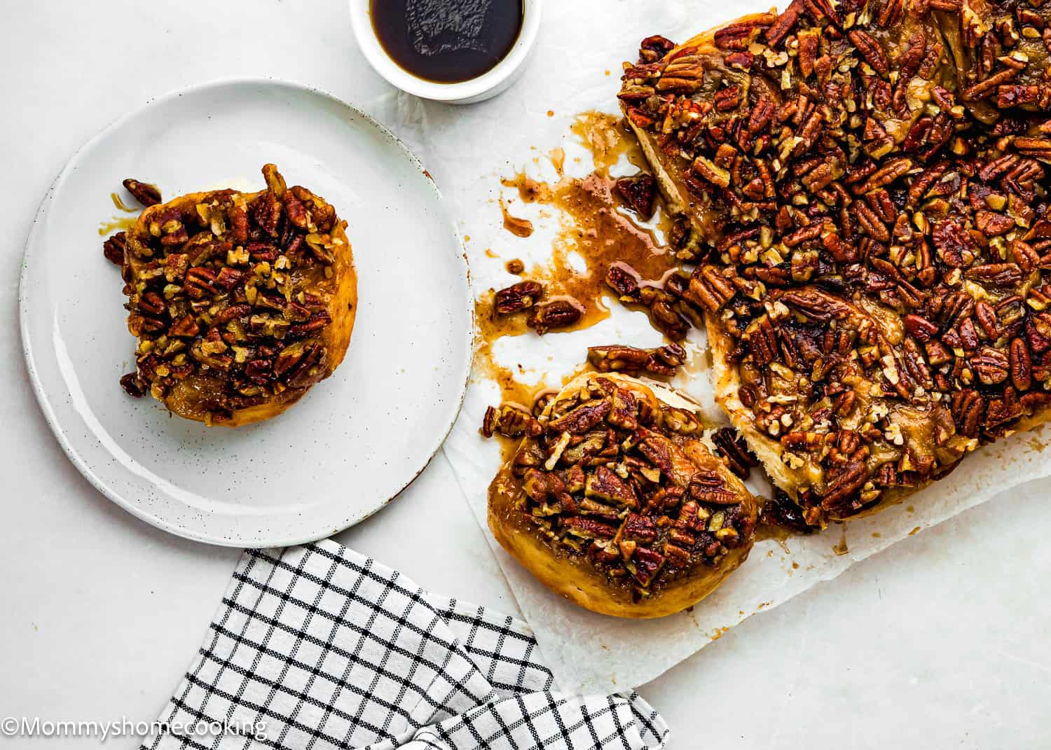 Eggless Sticky Buns, one served on a white plate, rest on a countertop with a cup of dark coffee and a checkered cloth nearby.