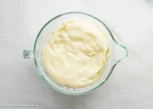 A glass bowl filled with risen Eggless Sticky Buns dough sits on a light-colored countertop.